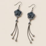 Midnight Blossom floral tassel earrings laid diagonally with deep indigo batik with pale flowing curves facing up and three chain tassels with rounded drops visible.