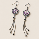 Lilac Serenade floral tassel earrings laid diagonally with lilac and pink petals with pale curved lines facing up and three chain tassels with rounded drops visible.
