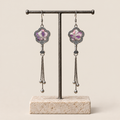 Lilac Serenade floral tassel earrings hanging from a T-bar stand, showing lilac and pink petals with pale curved lines and three chain tassels with rounded drops.