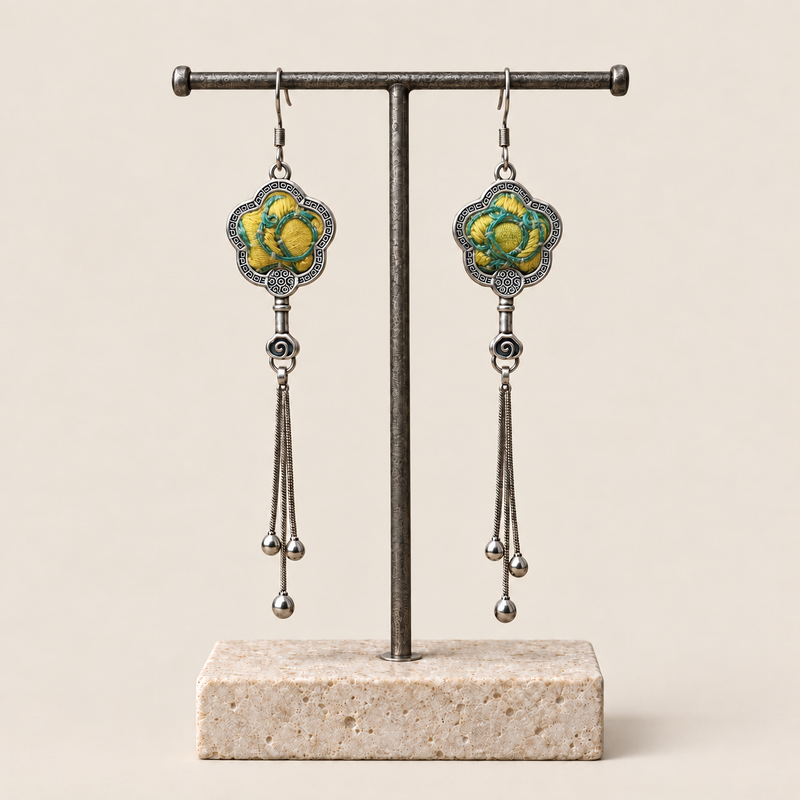 Golden Meadow floral tassel earrings hanging from a T-bar stand, showing yellow and green floral embroidery and three chain tassels with rounded drops.