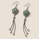 Jade Whisper floral tassel earrings laid diagonally with soft green floral embroidery facing up and three chain tassels with rounded drops visible.