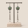 Jade Whisper floral tassel earrings hanging from a T-bar stand, showing soft green floral embroidery and three chain tassels with rounded drops.