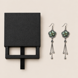 Black drawer-style box pictured beside Jade Whisper floral tassel earrings, with textile fronts and metal relief backs visible.
