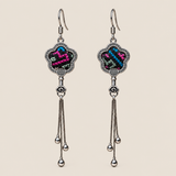 Rose Constellation floral tassel earrings, front view showing pink, green and blue geometric stitches on a dark ground in flower-shaped metal frames with three chain tassels with rounded drops.
