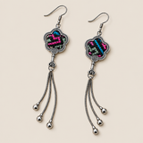 Rose Constellation floral tassel earrings laid diagonally with pink, green and blue geometric stitches on a dark ground facing up and three chain tassels with rounded drops visible.