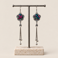 Rose Constellation floral tassel earrings hanging from a T-bar stand, showing pink, green and blue geometric stitches on a dark ground and three chain tassels with rounded drops.