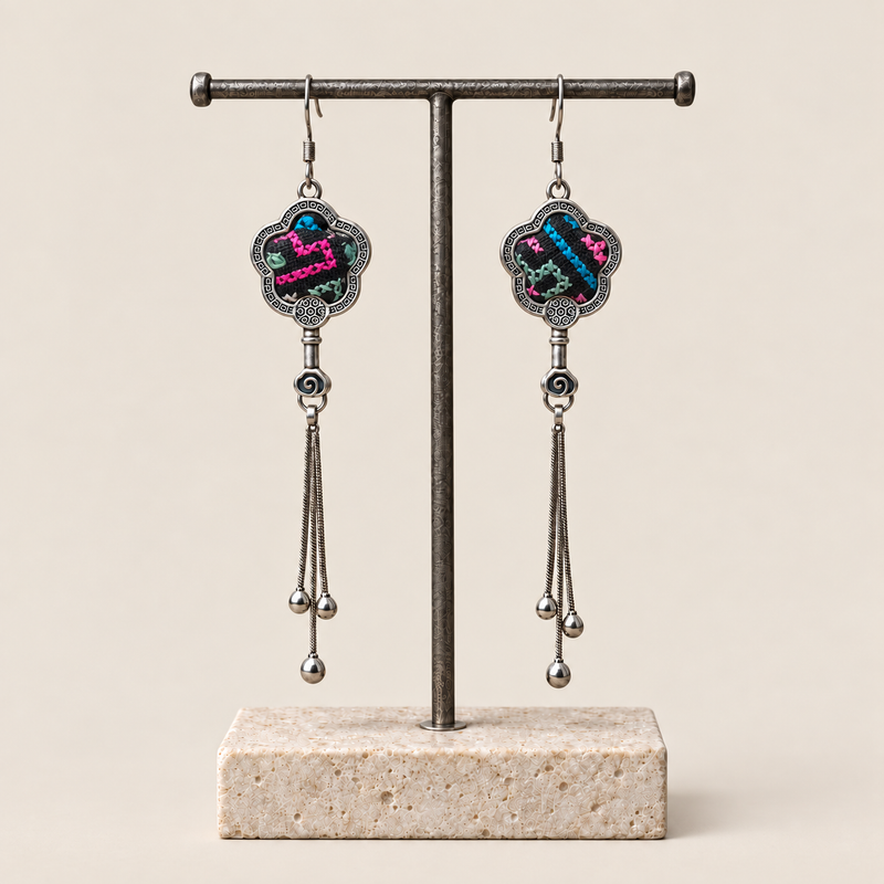 Rose Constellation floral tassel earrings hanging from a T-bar stand, showing pink, green and blue geometric stitches on a dark ground and three chain tassels with rounded drops.