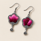 Fuchsia Dawn floral earrings laid diagonally with radiating fuchsia and rose-pink petals on a dark ground facing up and small spiral drops visible.