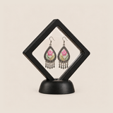 Blush Reverie floral earrings displayed inside a clear diamond-shaped frame on a black stand.