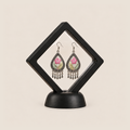 Blush Reverie floral earrings displayed inside a clear diamond-shaped frame on a black stand.