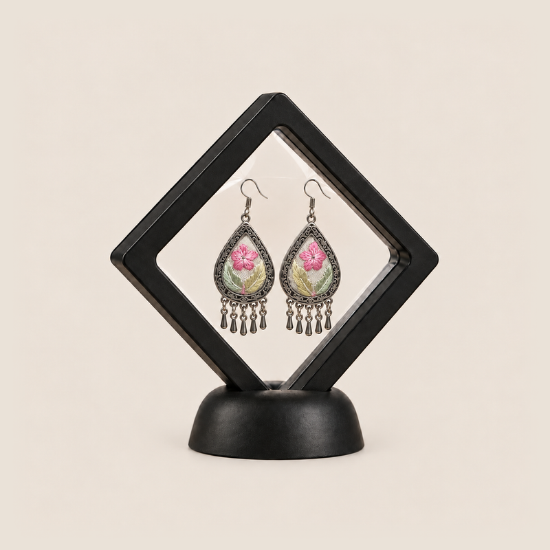 Blush Reverie floral earrings displayed inside a clear diamond-shaped frame on a black stand.