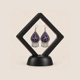 Violet Arabesque earrings displayed inside a clear diamond-shaped frame with a black border and base.