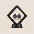 Violet Arabesque earrings displayed inside a clear diamond-shaped frame with a black border and base.