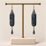 Indigo Stillness batik earrings hanging from a gold-tone display stand, with one engraved teardrop beneath each panel.