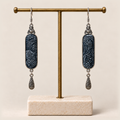Indigo Stillness batik earrings hanging from a gold-tone display stand, with one engraved teardrop beneath each panel.