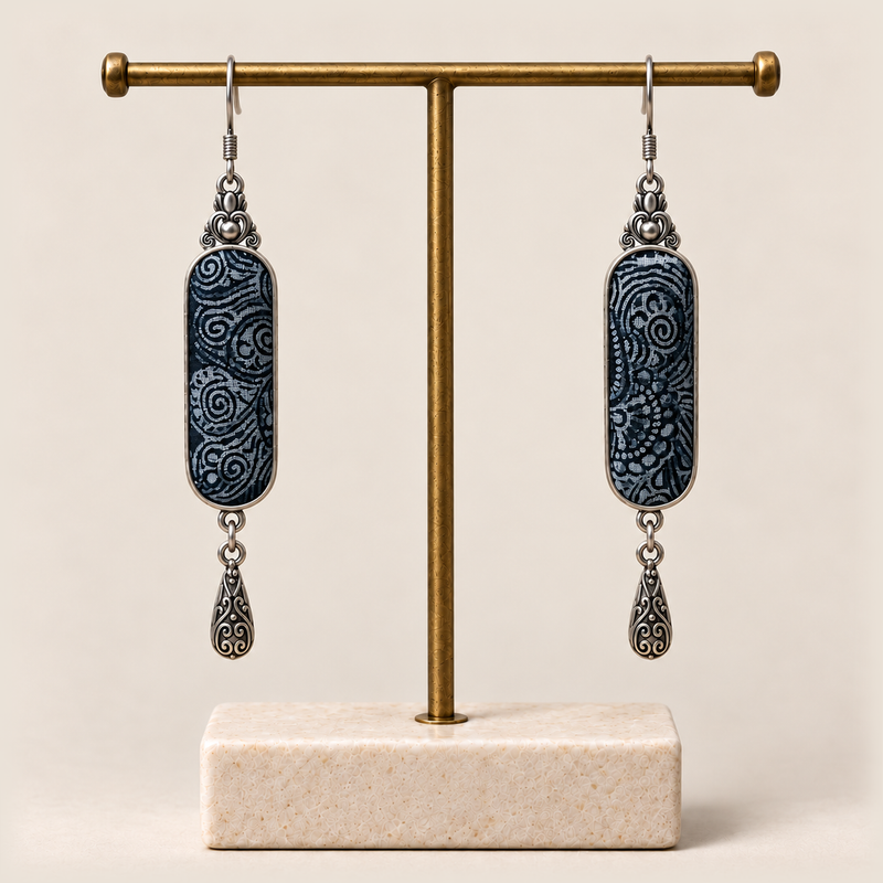 Indigo Stillness batik earrings hanging from a gold-tone display stand, with one engraved teardrop beneath each panel.