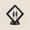 Pastel Cadence embroidered earrings inside a black diamond-shaped display frame on a matching base.