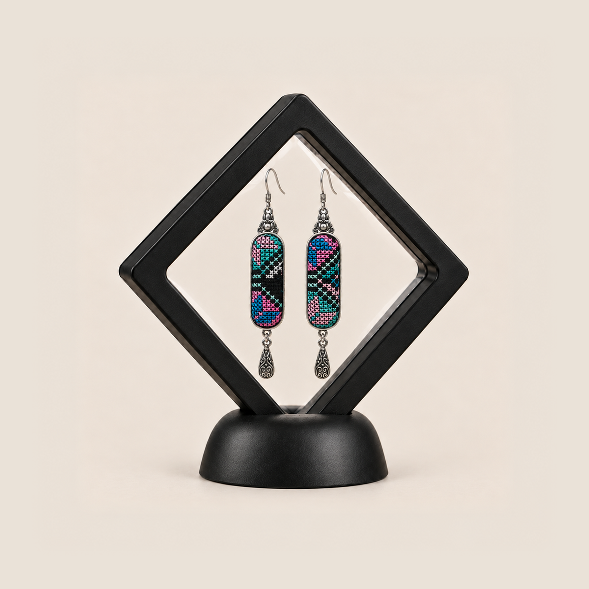Pastel Cadence embroidered earrings inside a black diamond-shaped display frame on a matching base.