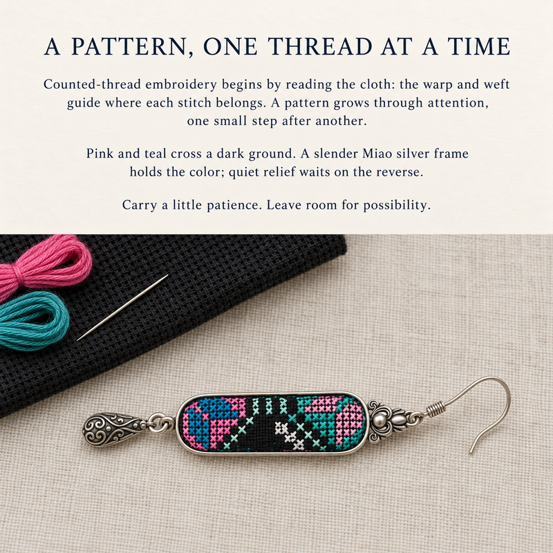 Counted-thread embroidery story card with a Pastel Cadence earring beside pink and teal thread, a needle and black cloth.