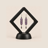 Violet Luminance earrings inside a black diamond-shaped display frame on a matching base.
