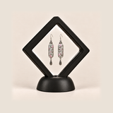 Pastel Arabesque earrings inside a black diamond-shaped display frame on a matching base.