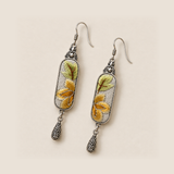Autumn Sonata earrings laid diagonally on a pale surface, showing green leaves above ochre petals on both embroidered panels.