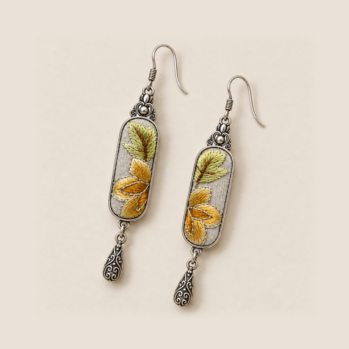 Autumn Sonata earrings laid diagonally on a pale surface, showing green leaves above ochre petals on both embroidered panels.