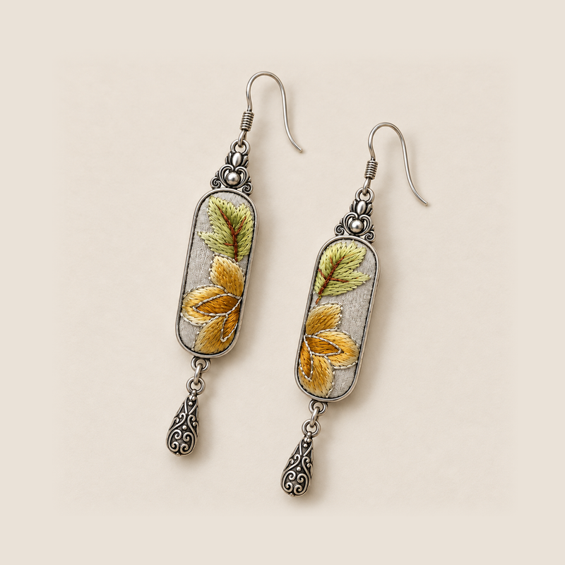 Autumn Sonata earrings laid diagonally on a pale surface, showing green leaves above ochre petals on both embroidered panels.