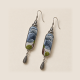 Blue Reverie earrings laid diagonally on a pale surface, showing layered blue-gray petals above green leaves.