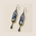 Blue Reverie earrings laid diagonally on a pale surface, showing layered blue-gray petals above green leaves.