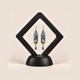 Blue Reverie earrings displayed inside a black diamond-shaped frame on a rounded black base.