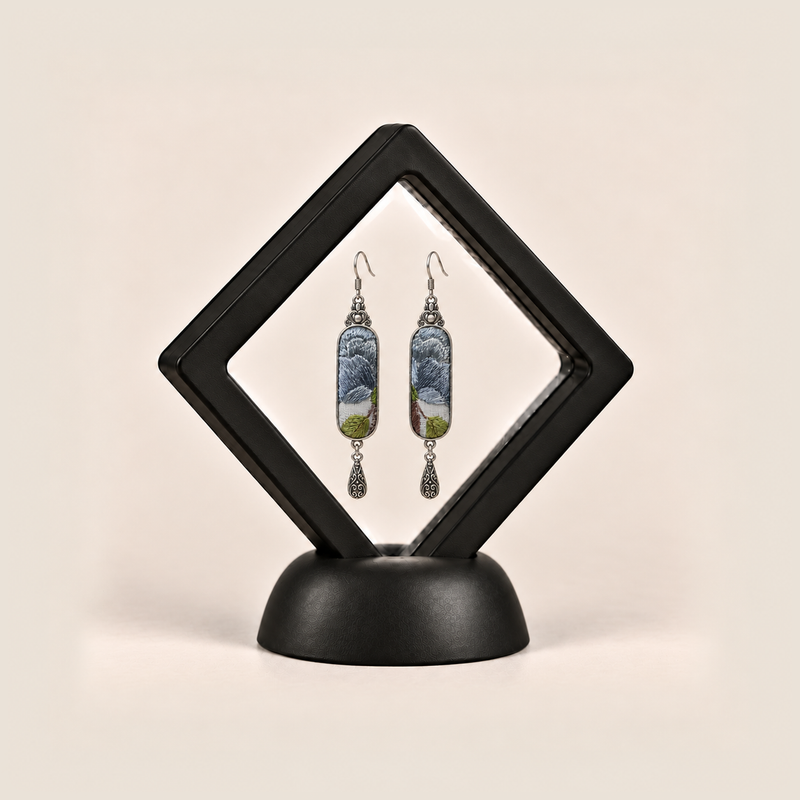 Blue Reverie earrings displayed inside a black diamond-shaped frame on a rounded black base.