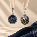 Indigo Cosmos earrings suspended from pale woven fabric strips, showing the indigo batik front and domed scrollwork reverse.