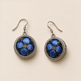 Sapphire Reverie earrings laid front-facing, showing blue petals and gold-colored accents on dark fabric within geometric circular rims.