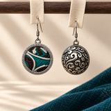 Jade Arabesque earrings hanging from pale woven strips over a wooden bar, showing the embroidered front and domed scrollwork reverse.