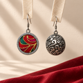Crimson Cadence earrings hanging from pale woven strips, showing the red embroidered front and domed scrollwork reverse above red cloth.