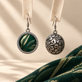 Forest Lustre earrings hanging from pale woven strips, showing the brocade front and domed scrollwork reverse above green patterned cloth.