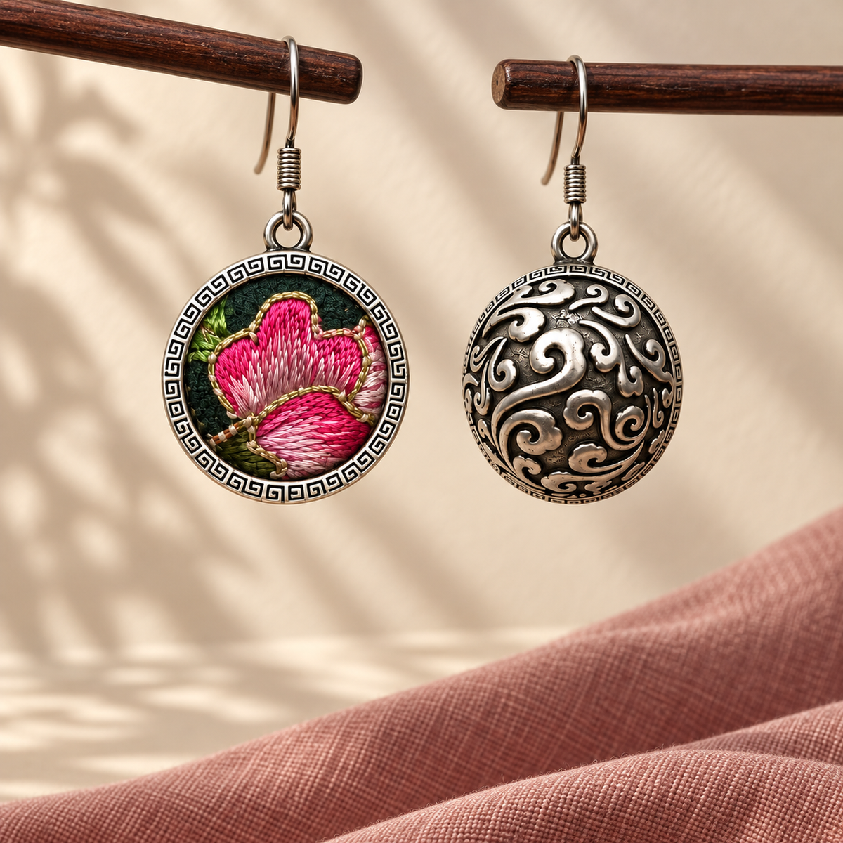 Rosy Aubade earrings hanging from wooden rods, showing the pink embroidered front and domed scrollwork reverse above pink cloth.