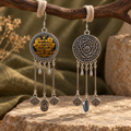 Golden Benediction earrings hanging from pale loops on a branch, showing the embroidered front, floral reverse and long rods with hanging charms.