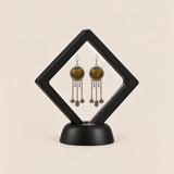 Golden Benediction earrings displayed inside a clear membrane in a black square frame set diagonally on a black base.