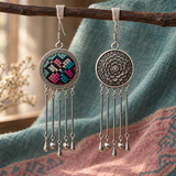 Pastel Refrain earrings suspended by pale fabric loops from a branch, one embroidered front and one floral reverse, against teal and pink woven cloth.