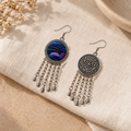 Violet Undertow earrings on cream woven fabric beside dried flowers and a ceramic dish, showing one blue-violet embroidered front and one floral reverse.