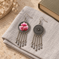 Blush Rhapsody earrings on folded pale woven fabric beside a book and dried flowers, showing one pink embroidered front and one floral metal reverse.
