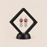 Blush Rhapsody pair displayed in a black square clear-membrane frame on a black stand, with both pink embroidered fronts facing forward.