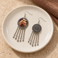 Garnet Serenade earrings on a cream ceramic plate beside brown woven fabric, showing one wine-red embroidered front and one floral metal reverse.
