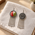 Coral Reverie earrings on folded cream woven cloth beside a pale vase, showing one coral-red and green embroidered front and one floral metal reverse.