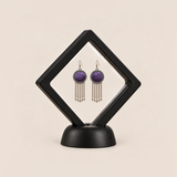 Violet Cadence pair displayed in a black square clear-membrane frame on a black stand, with both purple geometric embroidered fronts facing forward.