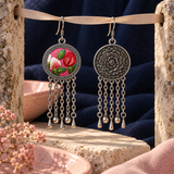 Petal Dialogue earrings hanging from a pale ribbon between stone supports, showing the embroidered front and floral reverse against navy fabric and pink ceramic props.