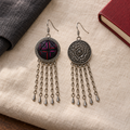 Crimson Confluence earrings on cream woven cloth beside dark fabric and a red book, showing one dark layered textile front and one floral metal reverse.