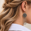 Model with wavy half-twisted hair wearing Golden Solace with the round floral metal reverse facing outward above five long chains with teardrop pendants.